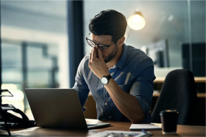 A man is sitting at his desk at work and rubbing his eyes under his glasses showing signs of stress. He is most likely feeling overwhelmed and consumed by work and is unable to find time to take care of his own personal needs.
