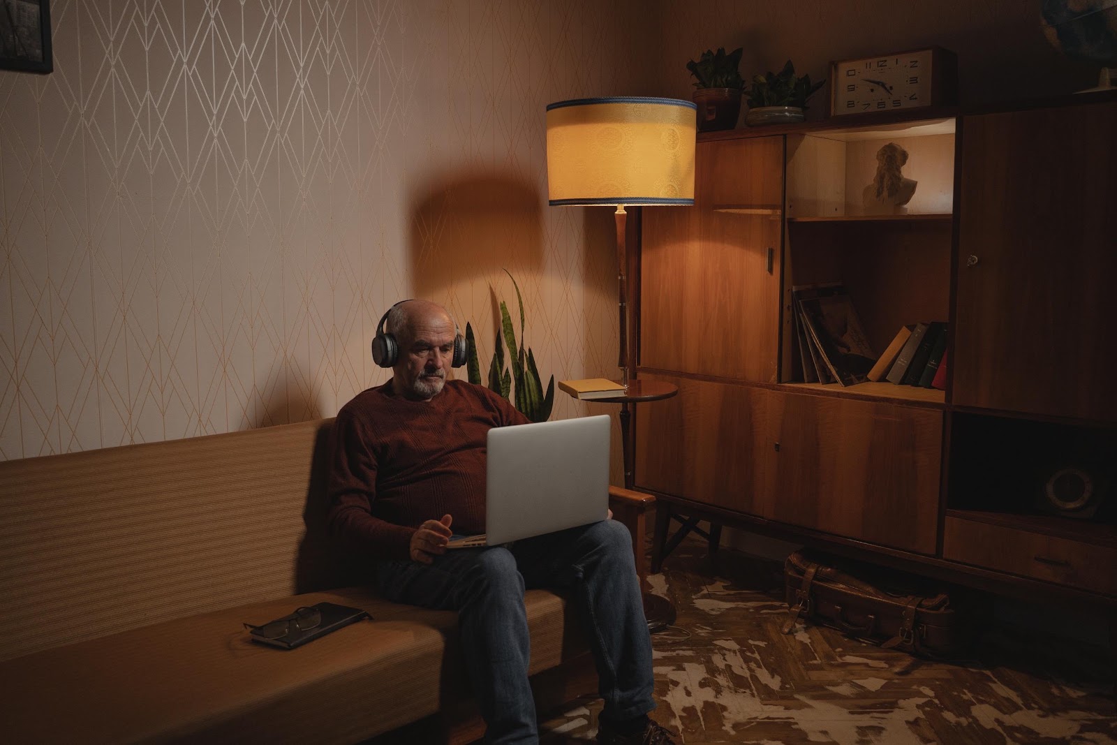 Older man sitting on a couch with headphones on and a laptop in his hands, reading about the benefits of online therapy for men.