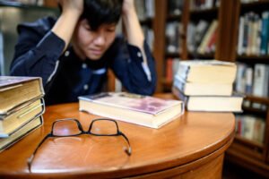Image of a man with his hands on his head staring at books. This represents someone who could benefit from meeting with an online therapist in California for decision making. Online therapy can help you take the next step forward.