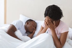 Image of a woman sitting next to her sleeping husband. Showing the pain that infidelity therapy in Portland, OR can help repair with support of an online therapist.
