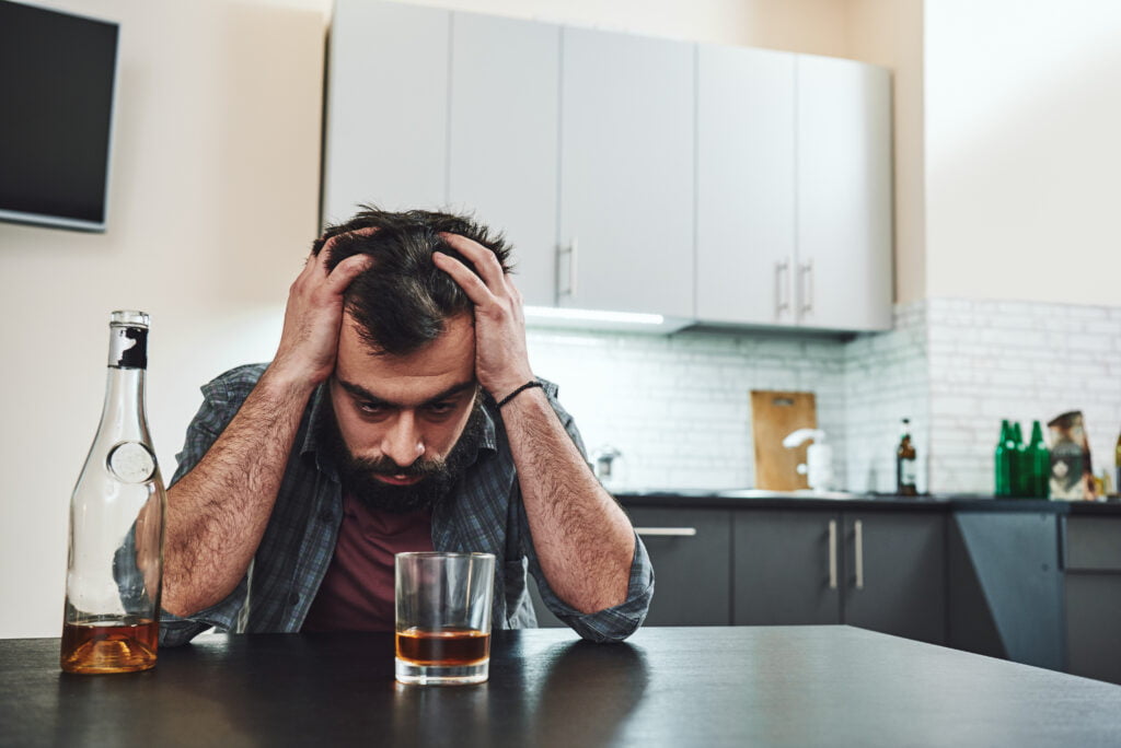 Drinking trouble. Depressed man sits at the table with his head in his hands. A glass of whiskey stands in front of him.