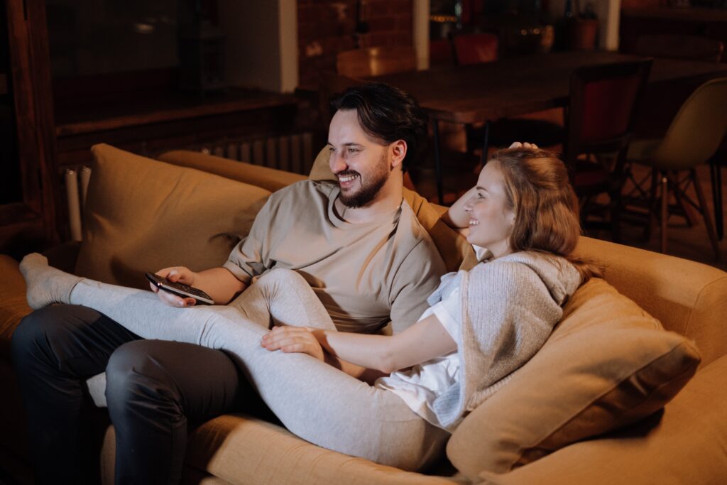 Couple sitting close on the couch, smiling, with the woman's legs on the man's, exhibiting physical intimacy and an ability to "turn towards" one another according to Gottman Method of Couples Therapy. 