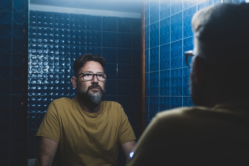 Portrait of sad and depressed man looking at himself in the mirror wondering how depression in men looks different than in women and if online therapy is the answer.