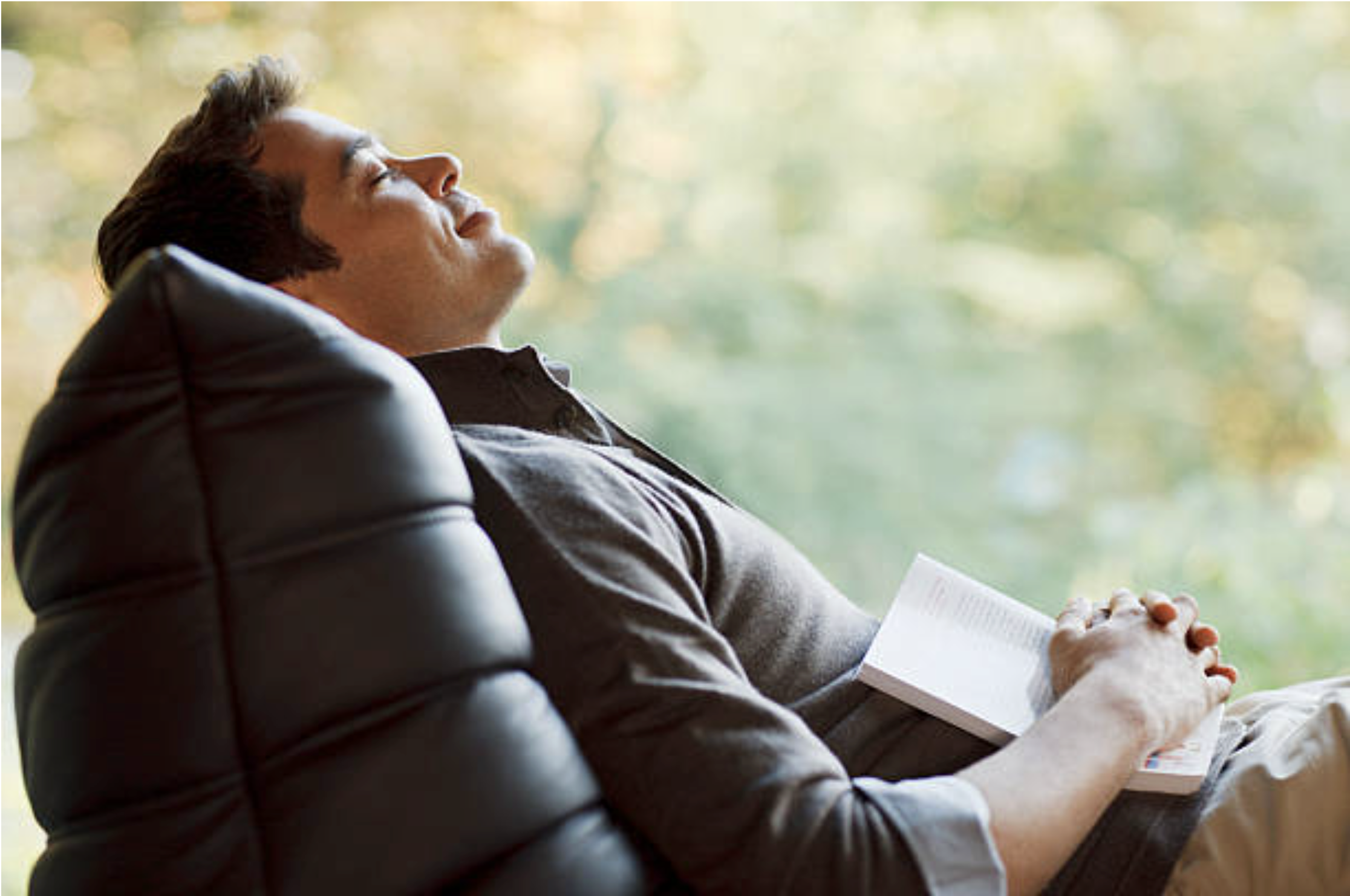 A man sitting back in a chair taking a break from reading a book to pause and breath and enjoy the time he has made for himself to do something that is relaxing.