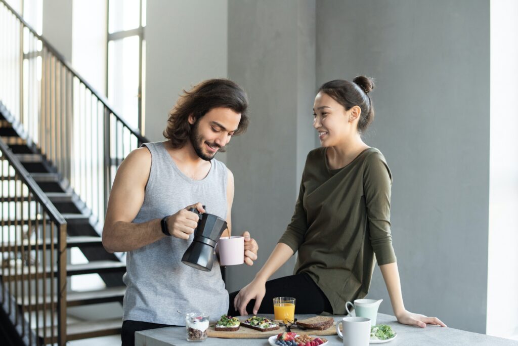 A married couple in the kitchen making coffee and breakfast together and smiling because they no longer feel like they're always fighting after couples therapy.