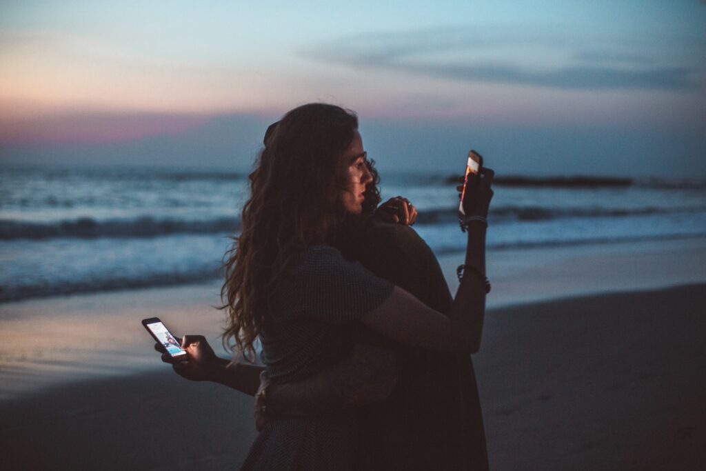 A couple hugging on the beach but rather than being present in the moment they are distracted by their cell phones behind each others back due to a lack of intimacy.