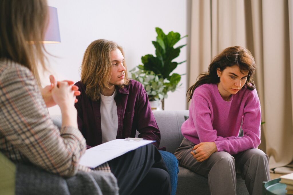 A man and woman sitting on a couch looking down, explaining to their therapist that they feel like they're always fighting and want to work through their issues.