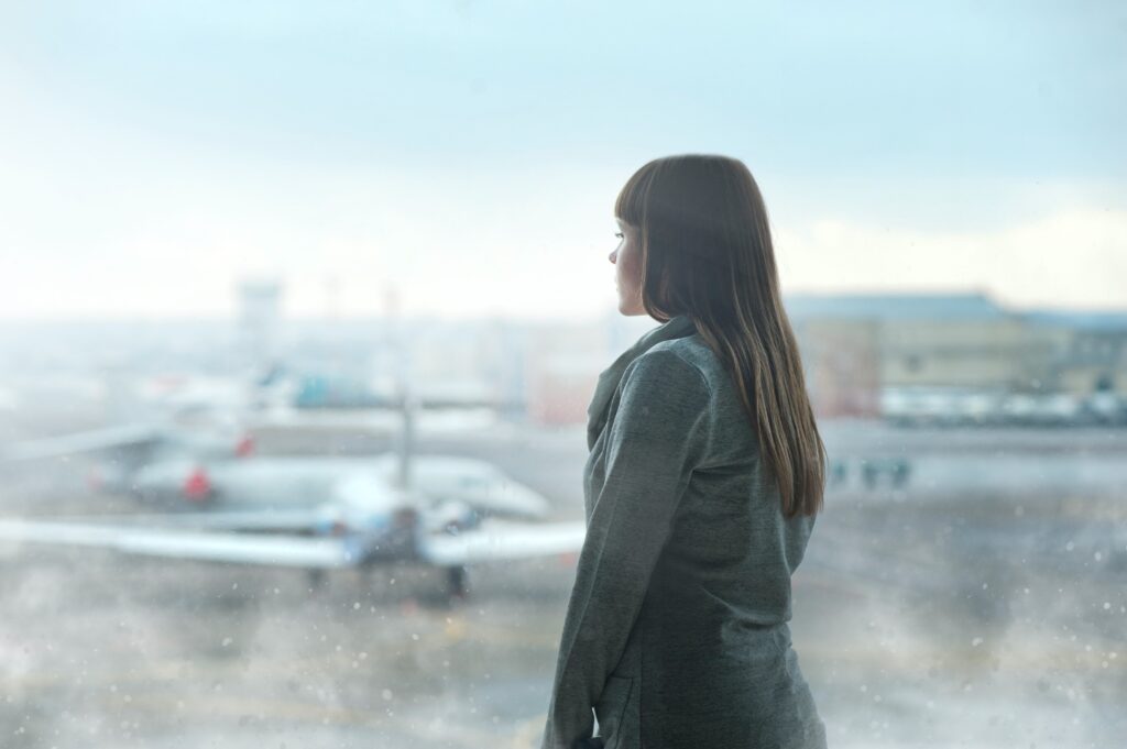 An image of a woman anxiously waiting inside the airport at her gate looking out the window at the airplanes on the tarmac