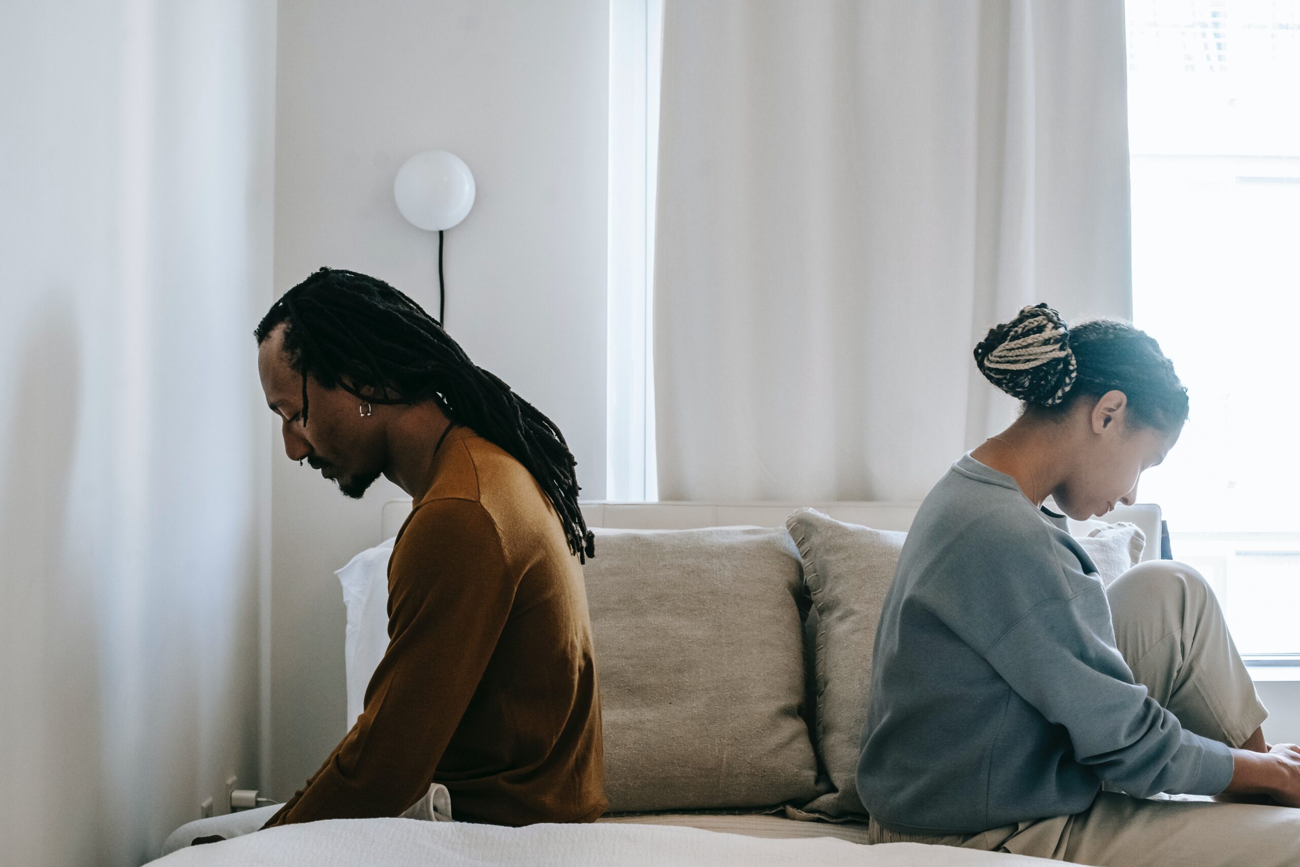 A disheartened couple sitting on opposite sides of a bed and facing away from each other as they face emotional turmoil due to infidelity.