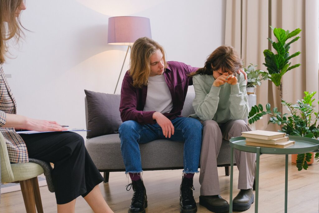 An image of a couple sitting on a couch in a therapists office, the woman is expressing emotional pain and the male is holding her.