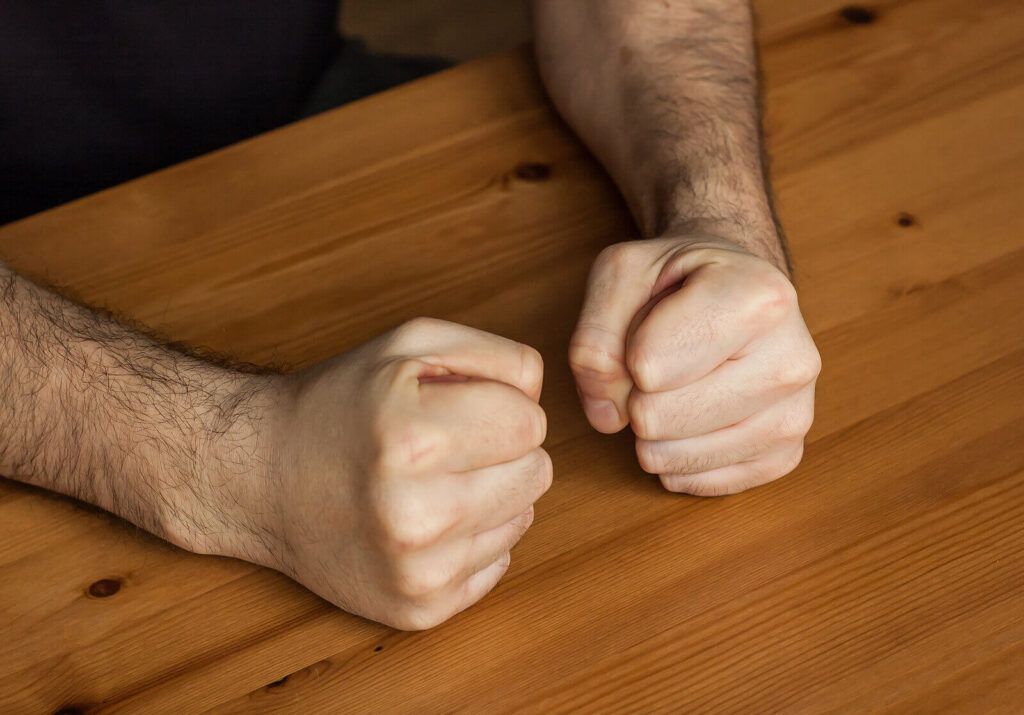 A close up of hands balled up in fists on a table. learn more about the support a San Diego therapist for men can offer in addressing men’s anger. learn more about men’s mental health in San Diego and other services today. 