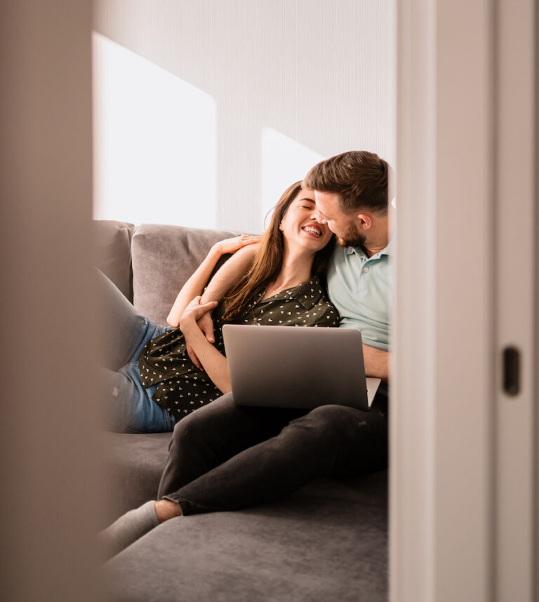 Smiling couple relaxing on the sofa with a notebook hugging each other, view between door. Represents successfully working on their relationship in couples counseling.