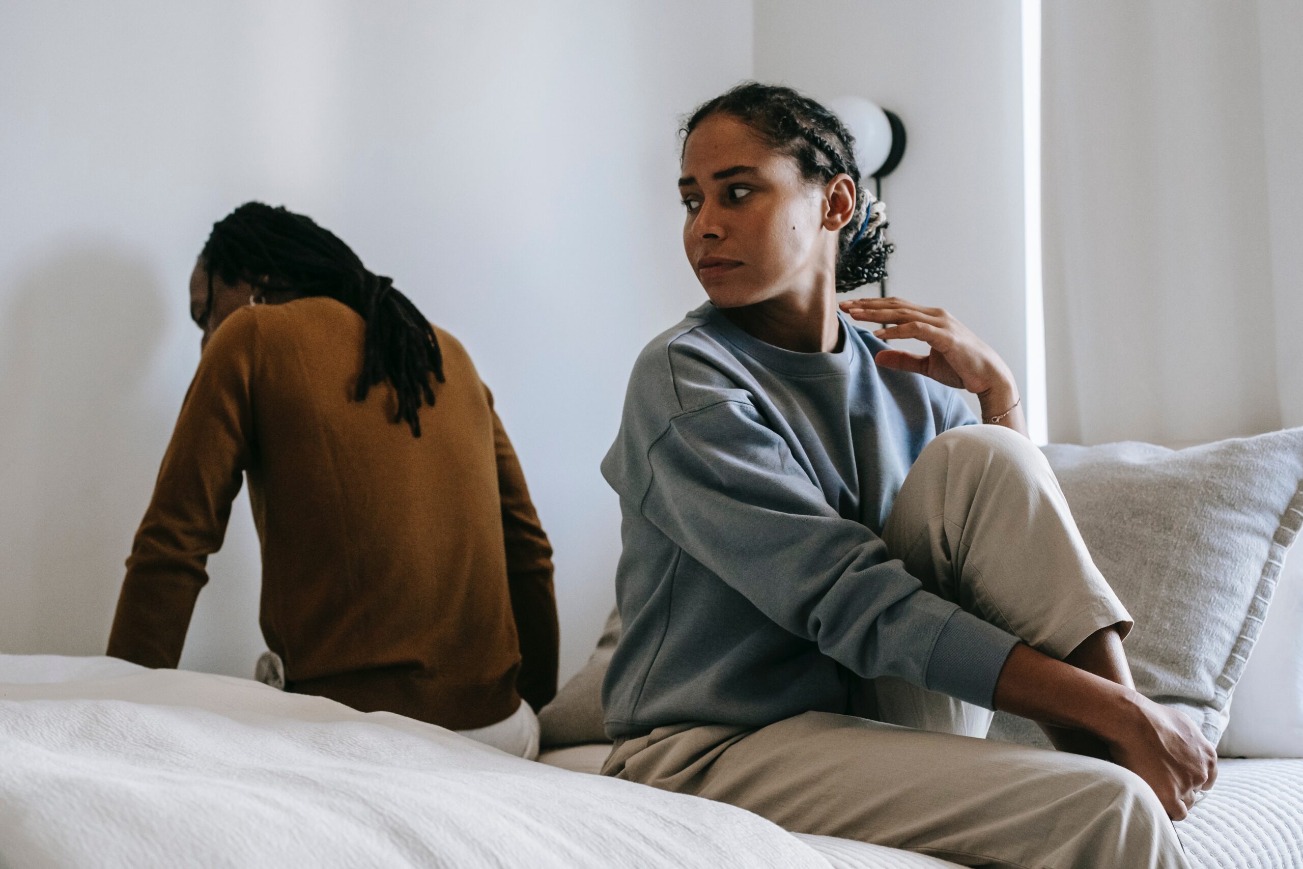 Image depicting a couple seated on a bed, each facing away from the other, showcasing a disconnect in their relationship. Their body language suggests tension and the need for couples therapy, as they appear distant and disconnected in their shared space.
