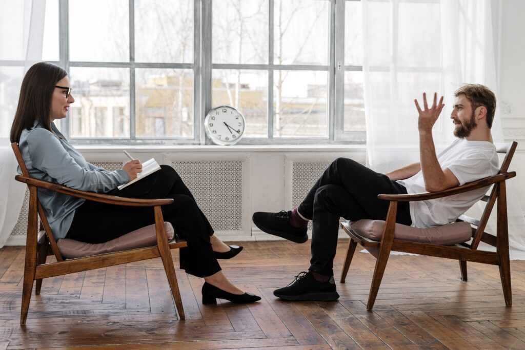 A man talks while sitting across from a woman holding a clipboard. Learn more about the importance of men’s mental health in San Diego and how online therapy for men in Portland, OR can help by contacting a San Diego therapist for men. 