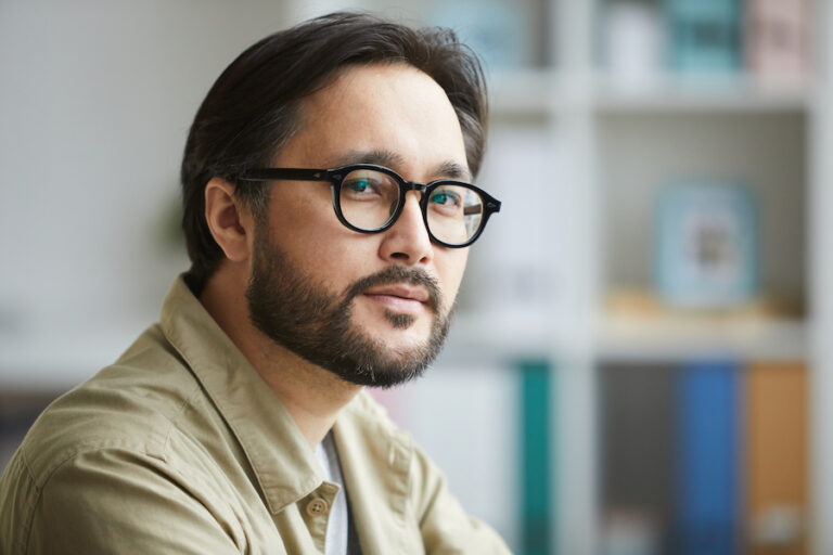 Portrait of confident young Asian man with beard and mustache wearing eyeglasses and shirt while working in office; feeling good about himself and his skills.