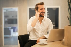 A man smiles while talking on a phone while in front of their laptop. This could represent the support offered when receiving anxiety help in San Diego. Search for anxiety help via an online therapist in California today.
