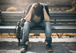 A man sits on a park bench with his head in his hands appearing distressed. Learn how a San Diego counselor can offer support via therapy by searching for online therapy in Oregon or "online therapist in California" today.
