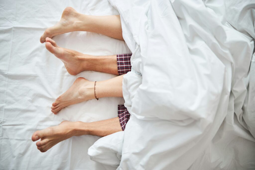 An image of a couples feet sticking out from under the covers in bed and struggling to find satisfaction in their sex life and in need of couples therapy in San Diego.