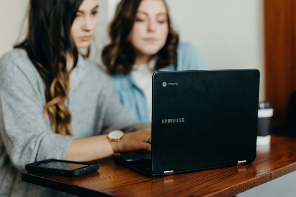 A woman checks her husband's social media profiles with her friend beside her, both focused on a laptop. This scene reflects relational trust issues and the need to control a partner, themes addressed by a Relational Life-Trained Therapist.