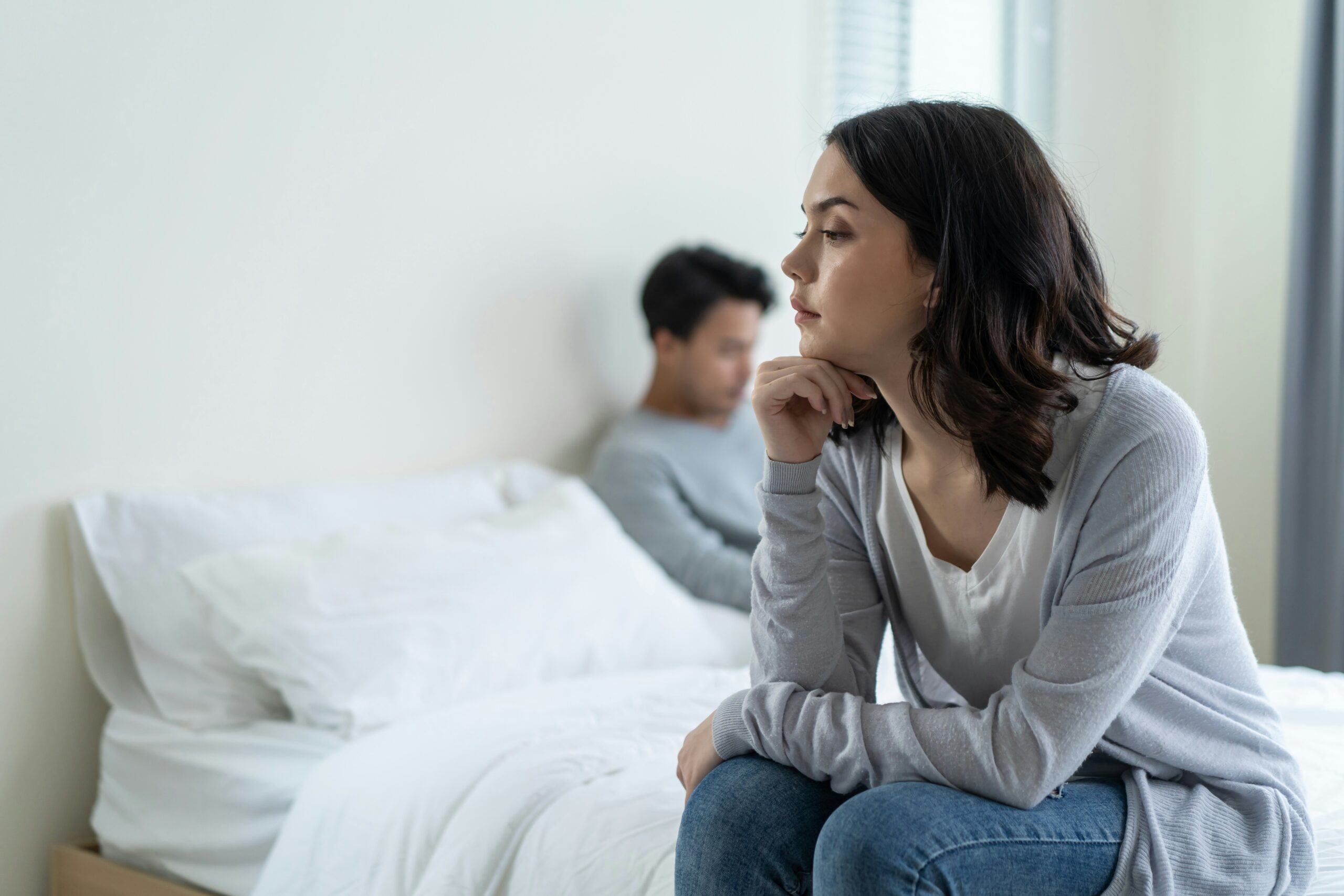 A woman sitting on the edge of a bed in deep thought, with her partner in the background. This visual highlights emotional distance, relational struggles, and the impact of needing to control a partner in relationships.