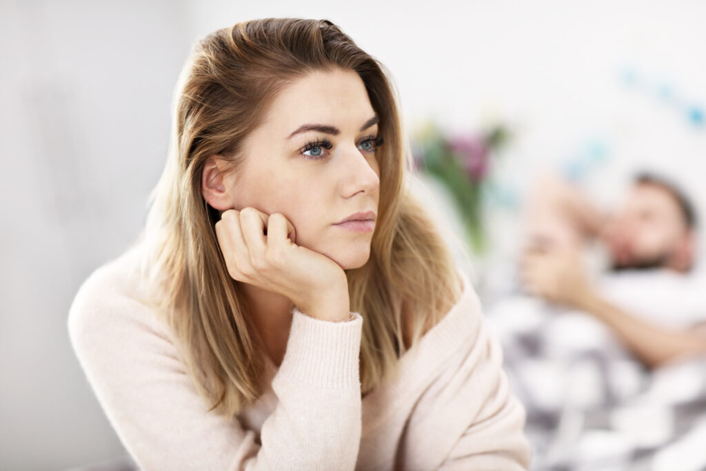 Concerned woman reflecting in foreground while distant partner lies in background, portraying relationship strain and the need for anxiety therapy for men.