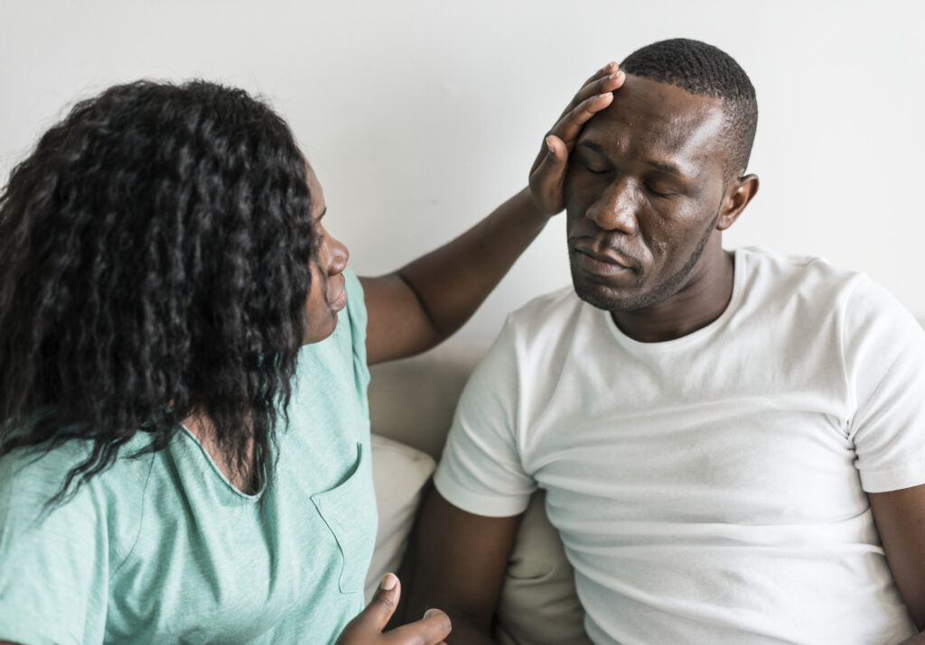 A worried woman gently touches her partner’s forehead as he sits with his eyes closed, visibly overwhelmed—illustrating the emotional weight of mental health struggles and the importance of therapists for men in San Diego.
