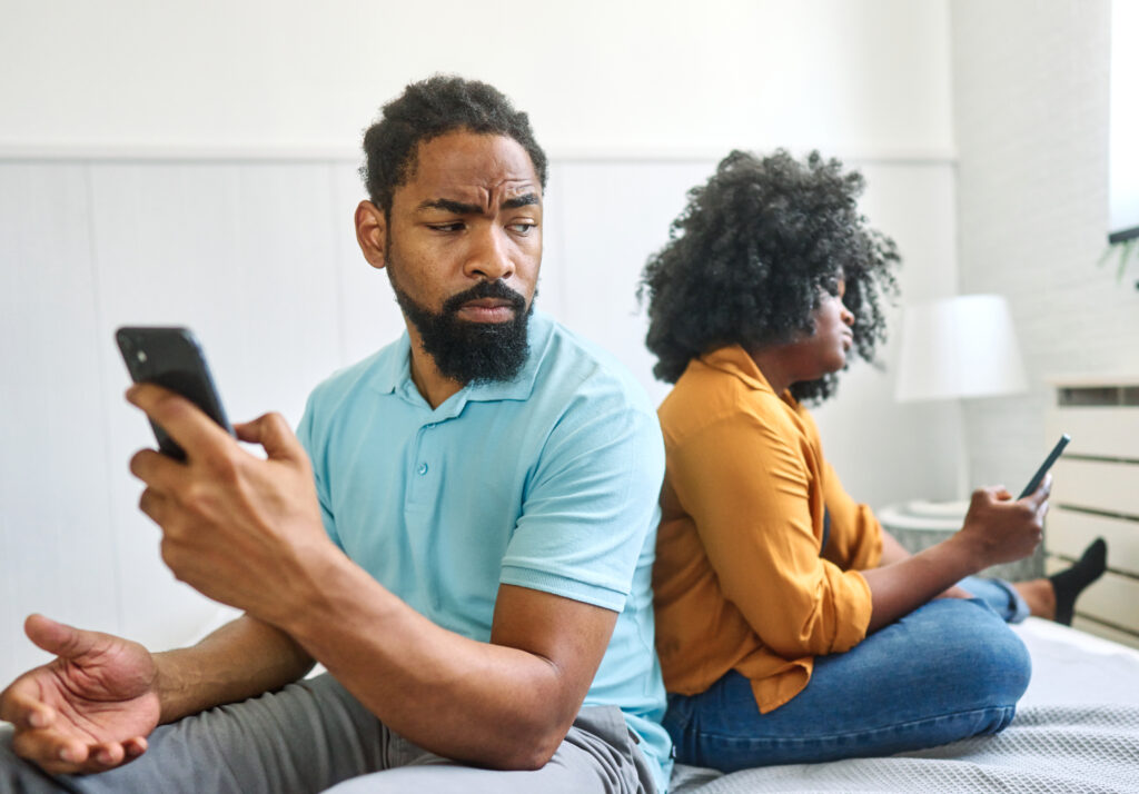 Frustrated couple sitting back-to-back on a bed, each holding a phone, showing disconnection and tension—an example of when to start couples therapy or seek relationship therapy in California.