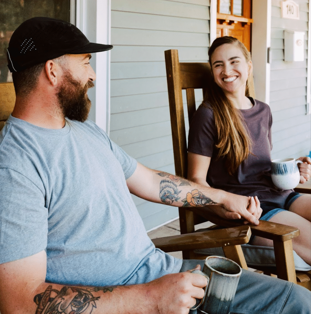 Smiling couple holding hands and coffee mugs on a porch, sharing a moment of connection—showing the positive impact of men’s mental health support.