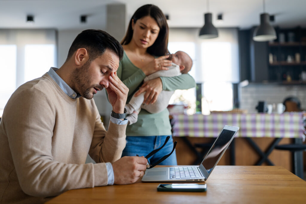 Stressed man sitting at a kitchen table with his hand on his face, laptop open, while his partner stands behind him holding a baby, looking concerned—representing the challenges of getting your partner to therapy and seeking men’s mental health support.