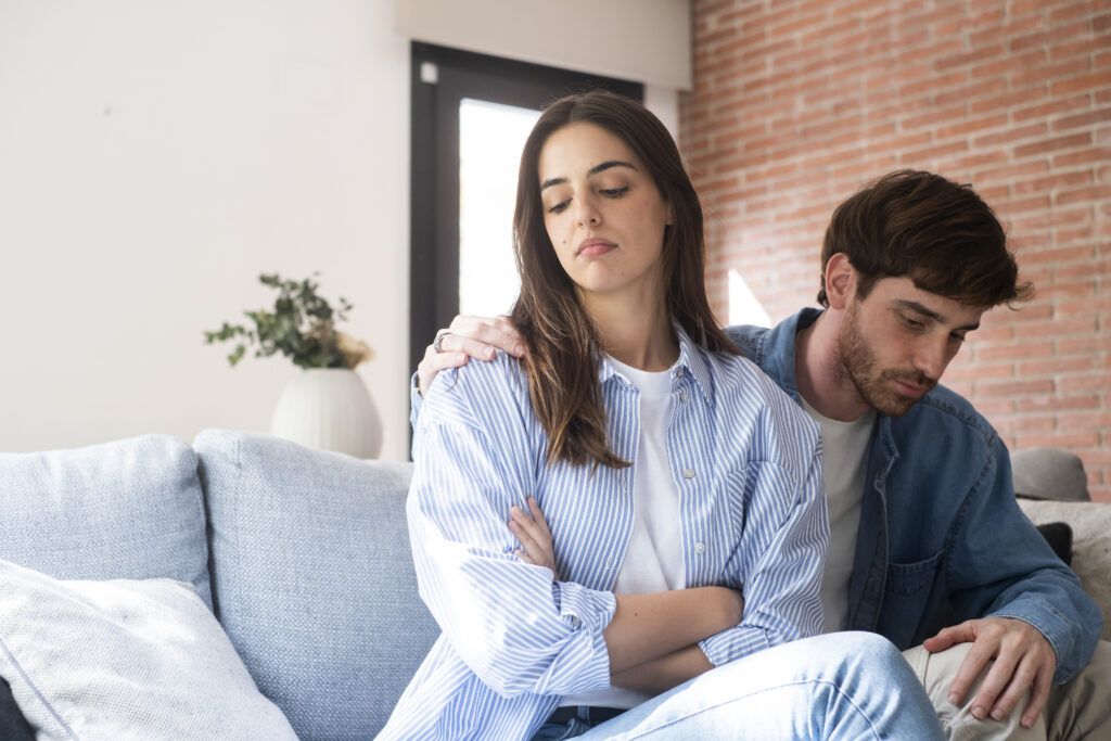 Unhappy couple sitting on a couch, man placing a hand on his partner’s shoulder while both look distant—illustrating struggles that couples counseling and relationship therapy can help address.