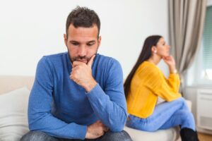 Worried man sitting in thought while his partner sits turned away behind him, highlighting common moments of conflict and the need for therapy for couples California.