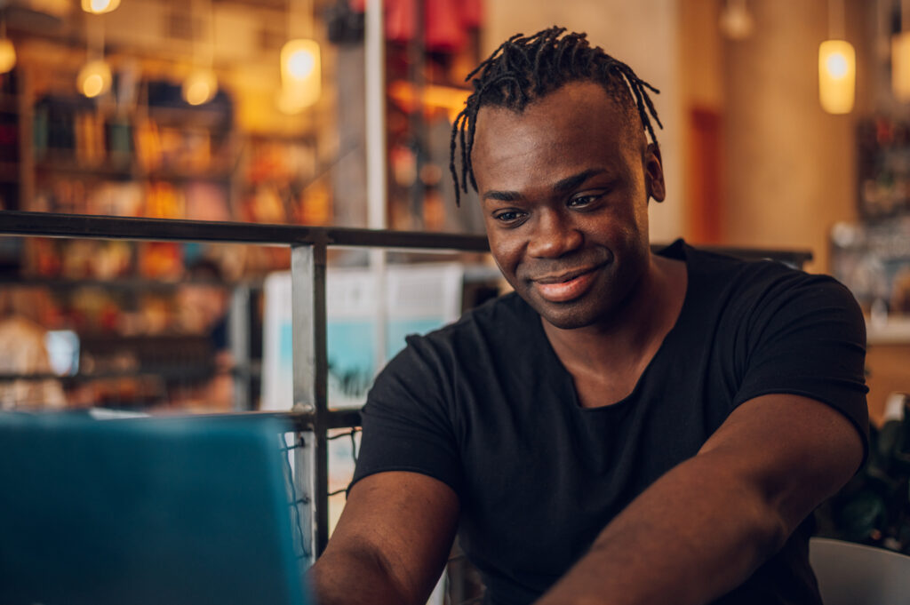 A man sitting in a café looking at his laptop with a relaxed smile, symbolizing the accessibility and comfort of online therapy in California and the growing benefits of virtual counseling for men.