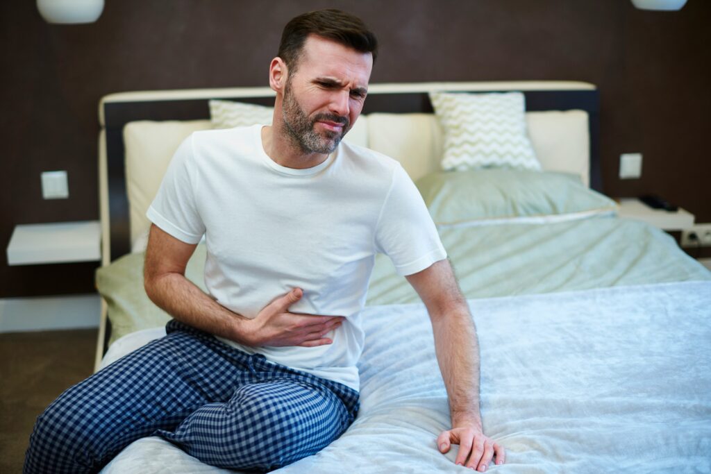 A man sitting on his bed clutching his stomach in discomfort, illustrating physical symptoms of hidden anxiety in men and the need for anxiety therapy in California to support men’s mental health.