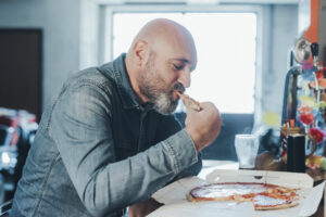 A man eating pizza alone at a table, representing hidden anxiety in men who use food or distraction to cope with stress; concept for anxiety therapy in California focused on men’s mental health.