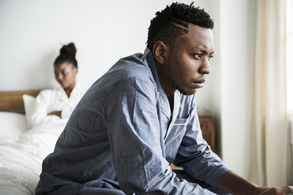A stressed man sits on the edge of the bed while his partner is in the background, reflecting relationship anxiety and how to stop overthinking in your relationship—Therapy for Anxiety & Relationships in San Diego, California with Stress Solutions.