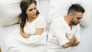 Couple lying in bed with the man turned away while the woman looks hurt and distant, illustrating disconnection explored in couples therapy for defensiveness in San Diego, CA.