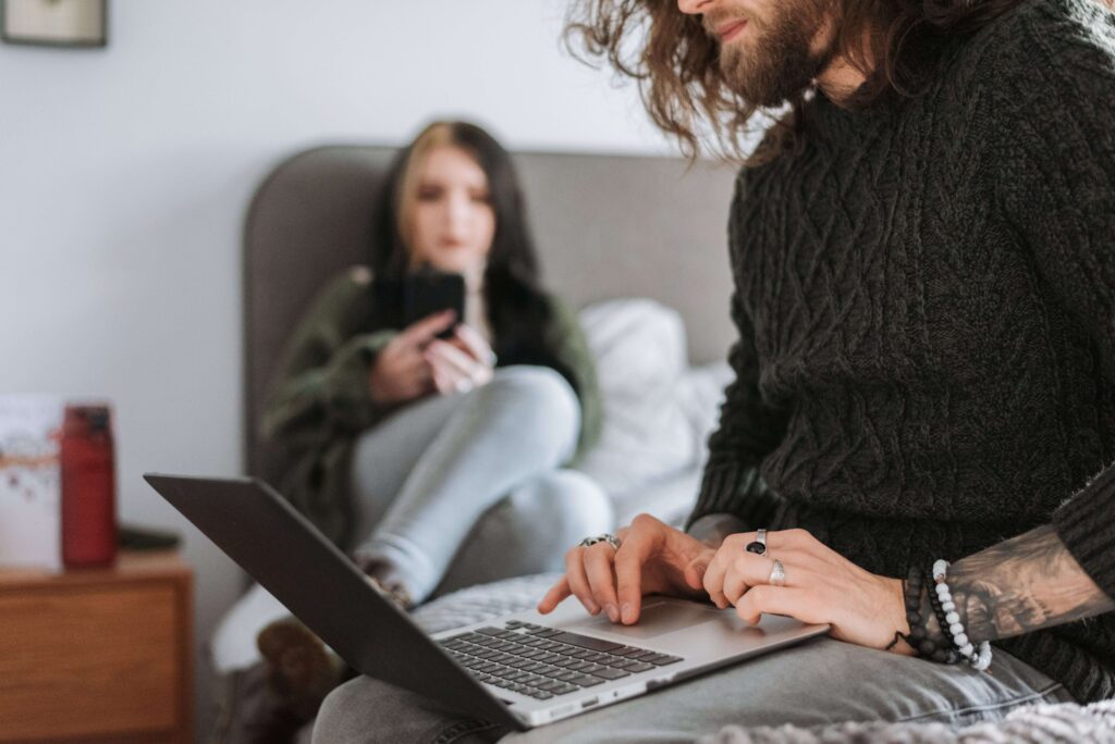 Man using a laptop while his partner sits nearby on a bed, illustrating the balance between togetherness and personal space supported by a relationship therapist in San Diego, CA.