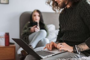 Man using a laptop while his partner sits nearby on a bed, illustrating the balance between togetherness and personal space supported by a relationship therapist in San Diego, CA.